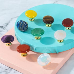 May include: A collection of gemstone cabinet knobs in various colors, including yellow, blue, brown, red, green, white, purple, and clear. Each knob has a gold-colored base. The knobs are arranged on a light blue and pink surface.