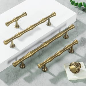 Puede incluir: Un conjunto de herrajes de gabinete de color latón antiguo, que incluye manijas y un pomo. Las manijas tienen un diseño inspirado en el bambú, con una forma cilíndrica y detalles curvos. Los herrajes se exhiben sobre una superficie blanca.