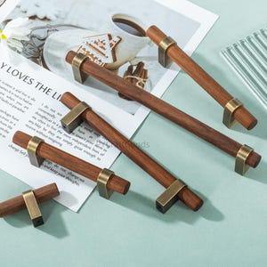 May include: A collection of wooden cabinet pulls with brass-colored accents. The pulls have a rectangular shape with a dark brown finish and are arranged on a light blue surface. The hardware is designed for furniture.