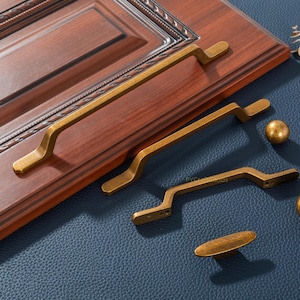 May include: A collection of antique brass cabinet hardware, including long bar pulls, curved pulls, a round knob, and an oval pull. The hardware is displayed on a dark wood cabinet door and a blue leather-textured surface.