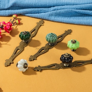 Peut inclure: Ensemble de poignées de meubles décoratives avec des plaques arrière en laiton antique. Les boutons en céramique sont de couleurs variées, dont vert, blanc et noir, avec des motifs côtelés ou floraux. Les poignées sont disposées sur une surface jaune.