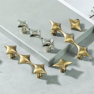 May include: A collection of decorative cabinet hardware in brass and silver finishes. The hardware includes star-shaped knobs and elongated pulls with a geometric design. The brand name "PYCraftGoods" is visible on the surface.