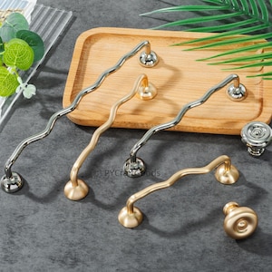 May include: A collection of decorative cabinet hardware, including wavy and knob-style pulls. The pulls are in silver and gold tones, with a modern design. The hardware is displayed on a wooden tray against a dark gray background.
