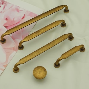 May include: An assortment of antique brass cabinet hardware, including four handles of varying lengths and a round knob. The hardware has a distressed finish, suggesting an aged appearance. The handles have curved ends and a slightly textured surface.