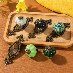 May include: A collection of decorative cabinet knobs in various colors, including white, black, green, and light green. Each knob has a unique, textured design and is mounted on an ornate, antique-style bronze base. The knobs are arranged on a wooden tray.
