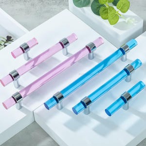Può includere: Maniglie per mobili rosa e blu, con un design cilindrico trasparente e dettagli in metallo argentato. L'hardware è esposto su una superficie bianca. Sullo sfondo sono visibili foglie verdi. Adatto a vari mobili.