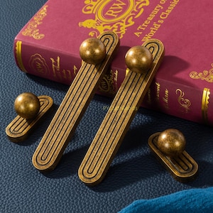May include: Four antique brass cabinet pulls and knobs. The pulls are long and rectangular with a ball at the top. The knobs are small and round. The hardware is displayed on a blue surface with a red book in the background.