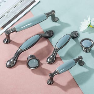 May include: A collection of cabinet hardware, including handles and knobs. The handles have a gunmetal finish with light blue ceramic inserts. The knobs are round with matching blue ceramic tops. The hardware is displayed on a pink and blue surface.