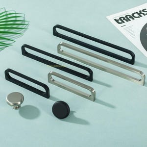 Op de afbeelding: Een verzameling kastbeslag, waaronder zwarte en zilverkleurige handgrepen en knoppen. De handgrepen zijn rechthoekig met afgeronde randen. Eén knop is zilver, de andere is zwart. De achtergrond is lichtblauw-groen.