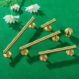 Peut inclure: Ensemble de quincaillerie de placard dorée, comprenant des poignées en forme de bambou et un bouton rond. L'ensemble est présenté sur un fond vert, avec des fleurs blanches. Les poignées sont de tailles variées, pour différentes dimensions de placards.