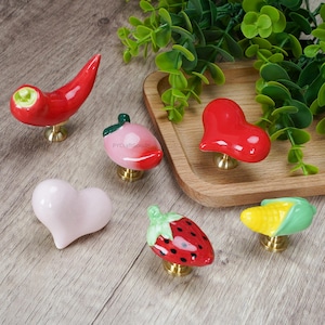 Könnte beinhalten: Eine Sammlung von Keramik-Schrankknöpfen in verschiedenen Obst- und Gemüseformen. Die Knöpfe umfassen eine rote Chilischote, eine rosa Erdbeere, ein rotes Herz, ein hellrosa Herz, eine rote Erdbeere und einen gelben Maiskolben. Jeder Knopf hat einen Messingfuß.