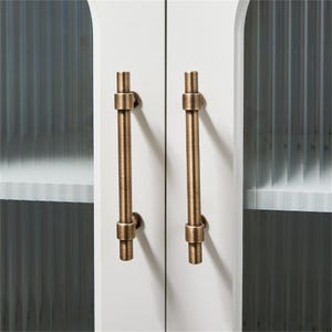 K&ouml;nnte beinhalten: Nahaufnahme eines wei&szlig;en Schranks mit zwei bronzefarbenen Stangengriffen. Die Griffe sind vertikal ausgerichtet und haben ein zylindrisches Design mit dekorativen Details. Die Schrankt&uuml;ren haben ein schlichtes, klares Design.