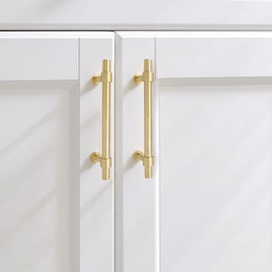 K&ouml;nnte beinhalten: Nahaufnahme von zwei wei&szlig;en Schrankt&uuml;ren mit goldfarbenen Stangengriffen. Die Griffe sind vertikal und T-f&ouml;rmig. Die T&uuml;ren haben ein sauberes, modernes Design. Die Griffe sind etwa 15 cm lang.