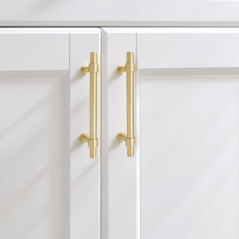 K&ouml;nnte beinhalten: Nahaufnahme von zwei wei&szlig;en Schrankt&uuml;ren mit goldfarbenen Stangengriffen. Die Griffe sind vertikal und T-f&ouml;rmig. Die T&uuml;ren haben ein sauberes, modernes Design. Die Griffe sind etwa 15 cm lang.