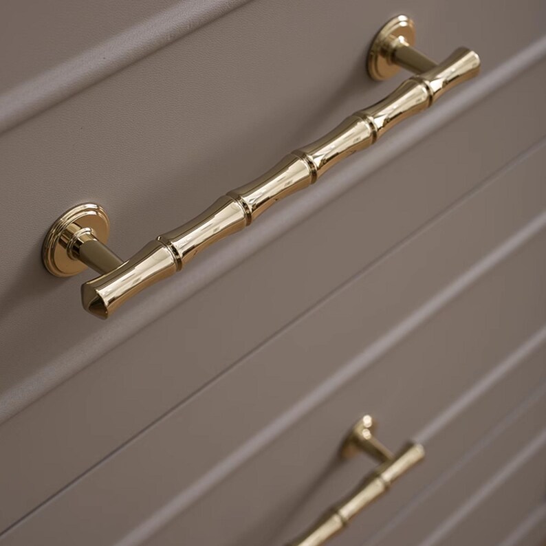 May include: Close-up of gold-coloured bamboo-style drawer pulls. The pulls are mounted on a light grey cabinet door with horizontal grooves. The hardware has a shiny, reflective finish.
