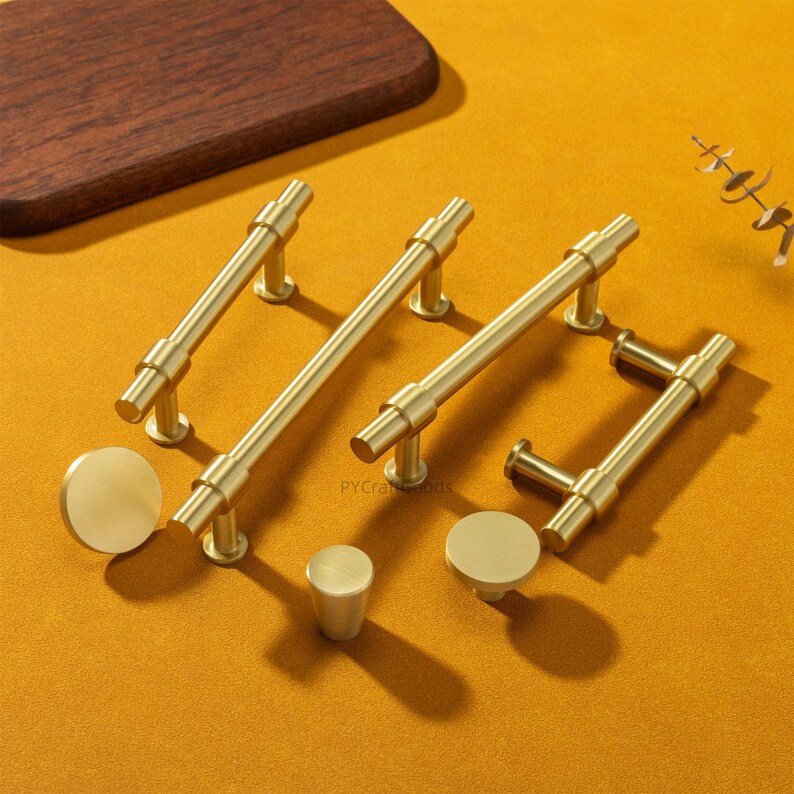 K&ouml;nnte beinhalten: Eine Sammlung goldfarbener Schrankbeschl&auml;ge, darunter Stangengriffe und runde Kn&ouml;pfe, angeordnet auf einer senffarbenen Oberfl&auml;che. Die Hardware hat ein schlankes, modernes Design mit zylindrischen Stangen und kreisf&ouml;rmigen oder zylindrischen Basen. Ein Holzschneidebrett befindet sich im Hintergrund.