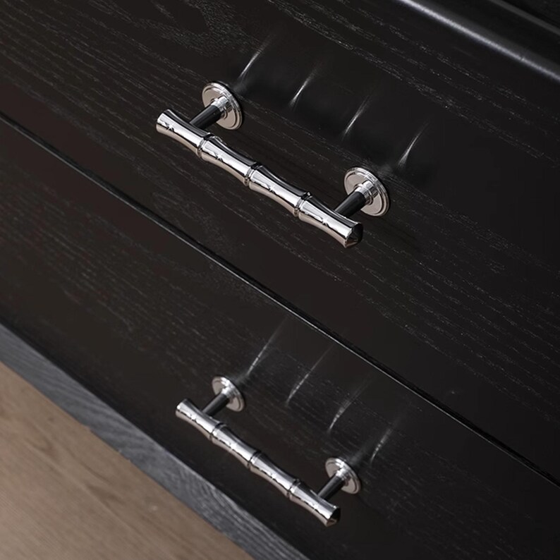 May include: Close-up of a black wooden chest of drawers with two drawers. Each drawer has a silver bamboo-style handle. The wood grain is visible, and the handles are mounted on circular silver plates.