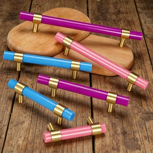 Puede incluir: Colección de tiradores de armario coloridos en tonos azul, rosa y morado. Cada tirador tiene un centro cilíndrico con detalles metálicos dorados y herrajes de montaje. Los tiradores están dispuestos sobre una superficie de madera.