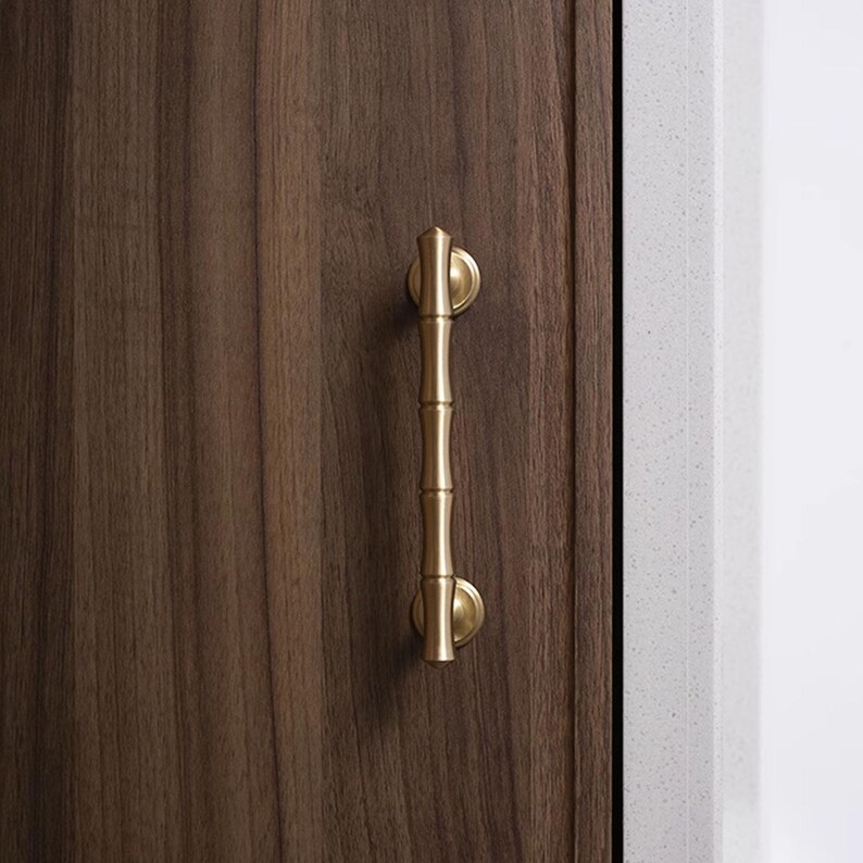 May include: A gold-coloured bamboo-style cabinet pull handle mounted on a dark brown wooden cabinet door. The handle has a vertical design with a cylindrical body and rounded ends.