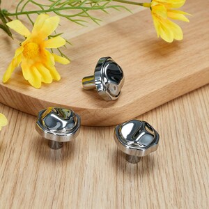 May include: Three polished silver cabinet knobs with a distinctive, textured surface. The knobs are displayed on a wooden board, with yellow flowers in the background. The knobs have a contemporary, minimalist design.