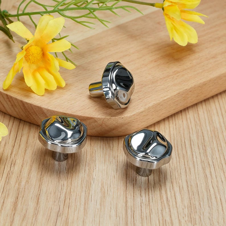 May include: Three polished silver cabinet knobs with a distinctive, textured surface. The knobs are displayed on a wooden board, with yellow flowers in the background. The knobs have a contemporary, minimalist design.