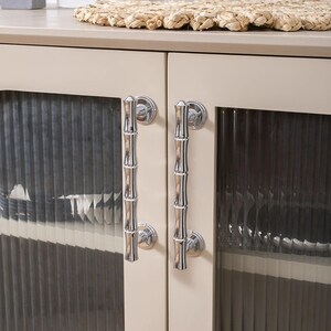 May include: A close-up of a cream-coloured cabinet with two glass doors. Each door has a polished silver bamboo-style handle. The glass doors have a vertical ribbed texture. A woven placemat sits on the cabinet top.