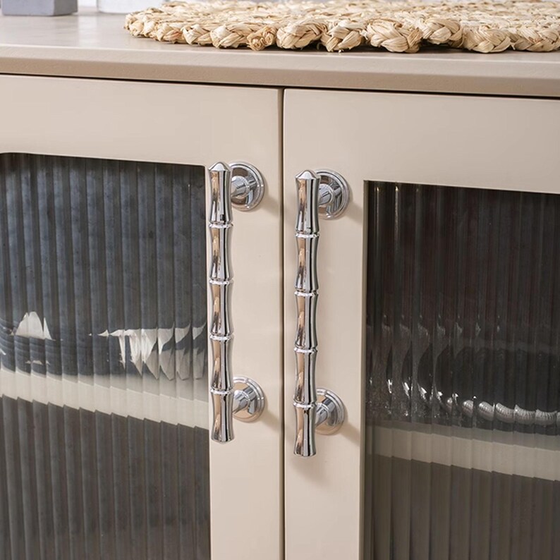 May include: A close-up of a cream-coloured cabinet with two glass doors. Each door has a polished silver bamboo-style handle. The glass doors have a vertical ribbed texture. A woven placemat sits on the cabinet top.