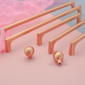 May include: A collection of rose gold cabinet hardware, including long bar pulls and round knobs. The pulls have a sleek, modern design with a curved base. The knobs are spherical. The hardware is displayed on a pink surface with a soft, blurred background.