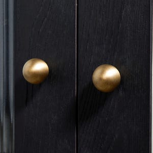 K&ouml;nnte beinhalten: Nahaufnahme einer schwarzen Holzschrankt&uuml;r mit zwei runden, messingfarbenen Kn&ouml;pfen. Die Kn&ouml;pfe sind auf den T&uuml;ren zentriert, die eine sichtbare Holzmaserung aufweisen. Der Schrank scheint Teil eines gr&ouml;&szlig;eren M&ouml;belst&uuml;cks zu sein.