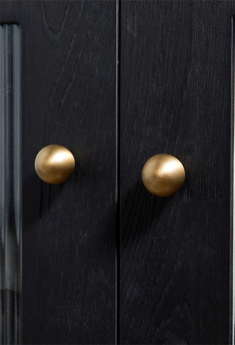 K&ouml;nnte beinhalten: Nahaufnahme einer schwarzen Holzschrankt&uuml;r mit zwei runden, messingfarbenen Kn&ouml;pfen. Die Kn&ouml;pfe sind auf den T&uuml;ren zentriert, die eine sichtbare Holzmaserung aufweisen. Der Schrank scheint Teil eines gr&ouml;&szlig;eren M&ouml;belst&uuml;cks zu sein.