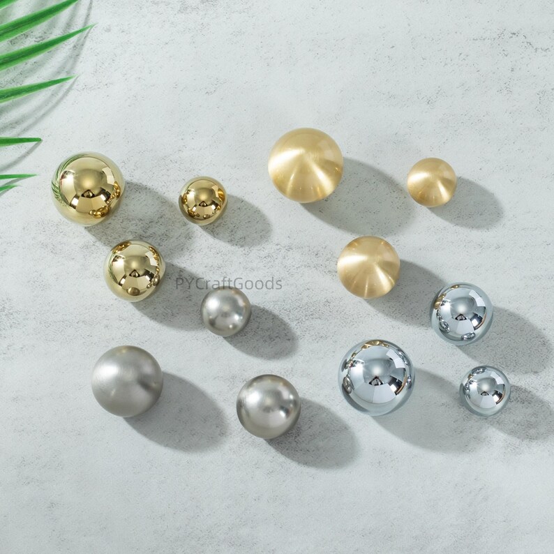 K&ouml;nnte beinhalten: Eine Auswahl an dekorativen Schrankkn&ouml;pfen in verschiedenen Gr&ouml;&szlig;en und Ausf&uuml;hrungen. Die Kn&ouml;pfe umfassen polierte goldene, geb&uuml;rstete Messing-, silberne und verchromte Kugeln. Die Kn&ouml;pfe sind auf einer hellgrauen Oberfl&auml;che angeordnet.