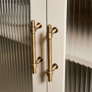 K&ouml;nnte beinhalten: Nahaufnahme einer Schrankt&uuml;r mit zwei vertikalen Griffen in antikem Messing. Die Schrankt&uuml;r hat eine geriffelte Glasscheibe. Die Griffe sind zylindrisch mit gebogener Halterung und rundem Sockel.