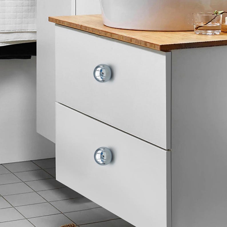 K&ouml;nnte beinhalten: Ein wei&szlig;er Badezimmerschrank mit zwei Schubladen, die jeweils mit einem gl&auml;nzenden, chromfarbenen Knauf versehen sind. Der Schrank hat eine hellfarbige Holzplatte, und ein wei&szlig;es Waschbecken ist dar&uuml;ber zu sehen. Die Kn&ouml;pfe verleihen einen modernen Touch.