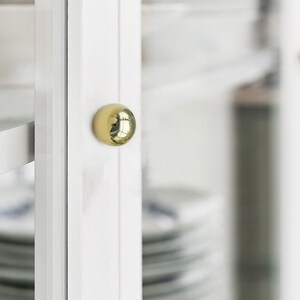 K&ouml;nnte beinhalten: Nahaufnahme einer wei&szlig;en Schrankt&uuml;r mit einem gl&auml;nzenden, goldfarbenen, kugelf&ouml;rmigen Knauf. Der Knauf spiegelt die Umgebung wider. Die Schrankt&uuml;r ist Teil eines gr&ouml;&szlig;eren wei&szlig;en Schranks, mit einem Blick auf Geschirr im Inneren.