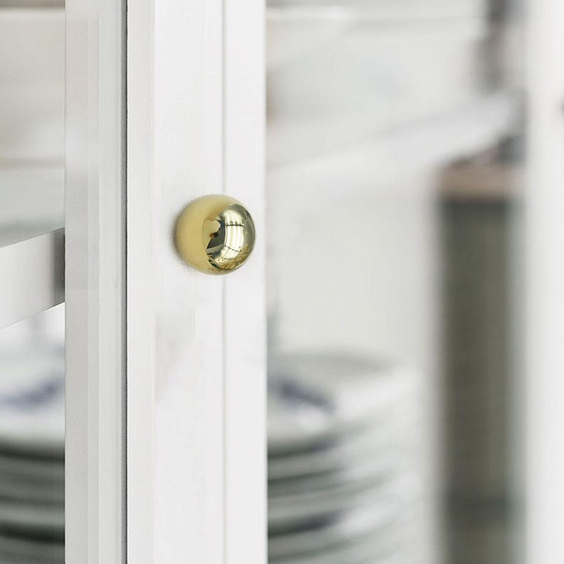 K&ouml;nnte beinhalten: Nahaufnahme einer wei&szlig;en Schrankt&uuml;r mit einem gl&auml;nzenden, goldfarbenen, kugelf&ouml;rmigen Knauf. Der Knauf spiegelt die Umgebung wider. Die Schrankt&uuml;r ist Teil eines gr&ouml;&szlig;eren wei&szlig;en Schranks, mit einem Blick auf Geschirr im Inneren.