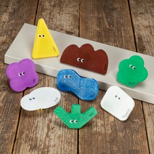 May include: A collection of eight colourful cabinet knobs in various shapes, including a yellow triangle, purple clover, brown cloud, blue blob, green cloud, white oval, green V-shape, and white square, each featuring two small eyes and a printed word.