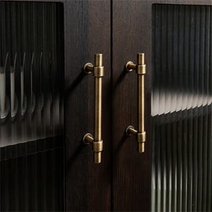 K&ouml;nnte beinhalten: Nahaufnahme eines dunklen Holzschranks mit zwei messingfarbenen Stangengriffen. Die Schrankt&uuml;ren haben eine strukturierte Glasscheibe. Die Griffe sind vertikal ausgerichtet und haben eine zylindrische Form.