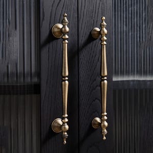 Peut inclure: Deux poign&eacute;es de porte en laiton antique avec des d&eacute;tails orn&eacute;s. Les poign&eacute;es sont verticales, avec une longue poign&eacute;e &eacute;lanc&eacute;e et des finials d&eacute;coratifs. Elles sont mont&eacute;es sur une porte en bois fonc&eacute;, cr&eacute;ant un look classique et &eacute;l&eacute;gant.