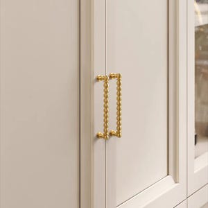 K&ouml;nnte beinhalten: Nahaufnahme einer wei&szlig;en Schrankt&uuml;r mit einem goldfarbenen Griff. Der Griff ist ein vertikaler Stab mit einer Reihe kleiner, runder Perlen. Die Schrankt&uuml;r ist Teil eines gr&ouml;&szlig;eren M&ouml;belst&uuml;cks.