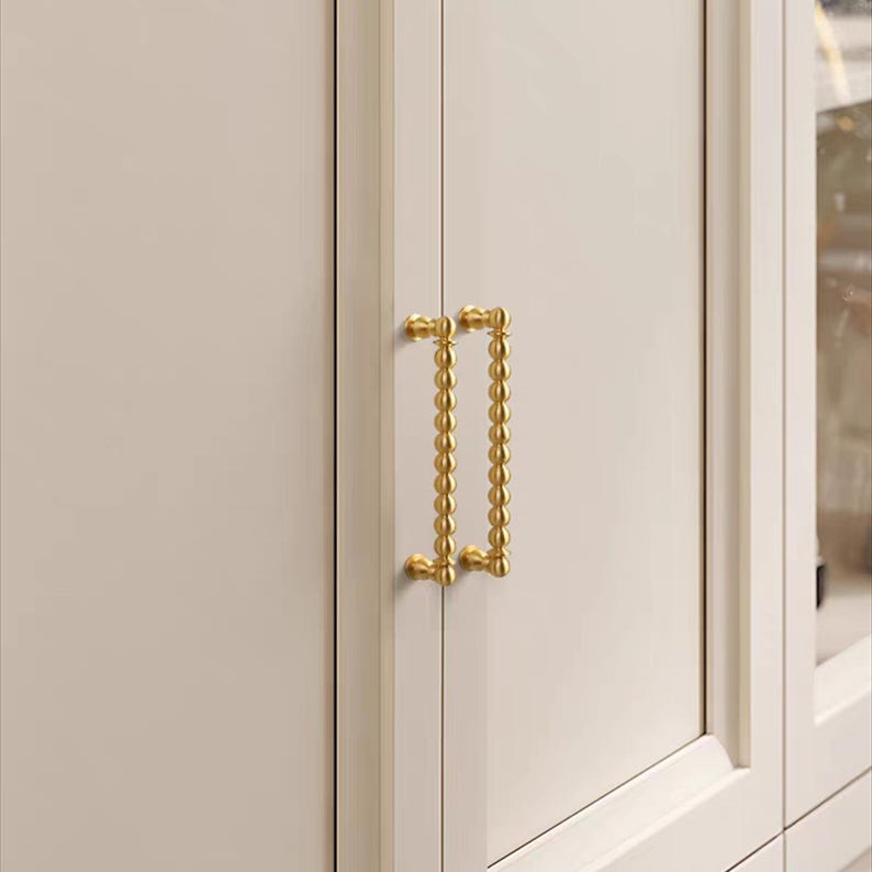 K&ouml;nnte beinhalten: Nahaufnahme einer wei&szlig;en Schrankt&uuml;r mit einem goldfarbenen Griff. Der Griff ist ein vertikaler Stab mit einer Reihe kleiner, runder Perlen. Die Schrankt&uuml;r ist Teil eines gr&ouml;&szlig;eren M&ouml;belst&uuml;cks.