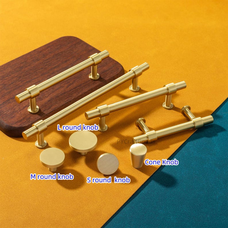 K&ouml;nnte beinhalten: Eine Auswahl an goldfarbenen Schrankbeschl&auml;gen, darunter verschiedene Gr&ouml;&szlig;en von runden Kn&ouml;pfen und Stangengriffen. Das Bild zeigt verschiedene Stile mit den Bezeichnungen "L round knob", "M round knob", "S round knob" und "Cone Knob". Die Beschl&auml;ge sind auf einem Holzbrett und einer farbigen Oberfl&auml;che angeordnet.