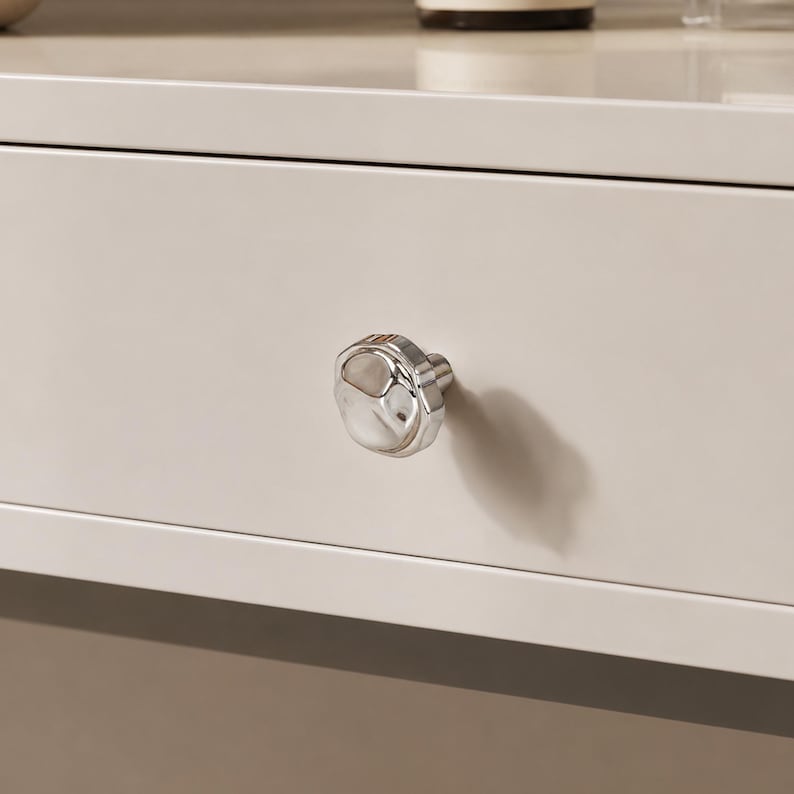 May include: Close-up of a light beige drawer with a silver, faceted knob. The knob has a rounded, irregular shape and reflects the light. The drawer is part of a larger piece of furniture.