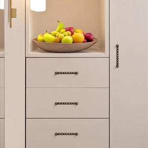 K&ouml;nnte beinhalten: Eine Holzschale mit verschiedenen Fr&uuml;chten, darunter &Auml;pfel, Birnen und Orangen, steht auf einem Regal &uuml;ber Schubladen. Die Schubladen haben dekorative, dunkel gef&auml;rbte Beschl&auml;ge. Das Wort "youke" ist oben links sichtbar.