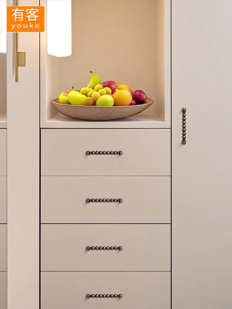 K&ouml;nnte beinhalten: Eine Holzschale mit verschiedenen Fr&uuml;chten, darunter &Auml;pfel, Birnen und Orangen, steht auf einem Regal &uuml;ber Schubladen. Die Schubladen haben dekorative, dunkel gef&auml;rbte Beschl&auml;ge. Das Wort "youke" ist oben links sichtbar.