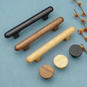 Op de afbeelding: Een verzameling houten kastbeslag, waaronder drie handgrepen in zwart, bruin en lichtbruin, en drie ronde knoppen. Het beslag wordt getoond op een blauw oppervlak, met een takje bloemen op de achtergrond.