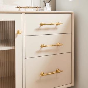 Peut inclure: Un meuble cr&egrave;me avec trois tiroirs et une porte vitr&eacute;e. Le meuble est orn&eacute; de poign&eacute;es dor&eacute;es sur les tiroirs et d'un bouton assorti sur la porte. Il s'agit d'un meuble.