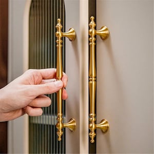 Peut inclure: Poign&eacute;es de porte de placard dor&eacute;es avec un design orn&eacute;. Les poign&eacute;es sont longues et fines, avec des d&eacute;tails d&eacute;coratifs et une esth&eacute;tique classique. Les poign&eacute;es sont mont&eacute;es sur une porte de placard gris clair.