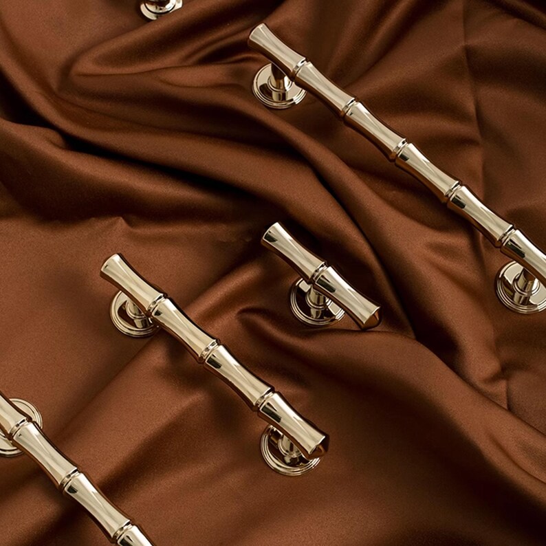 May include: Close-up of several gold-coloured cabinet handles with a bamboo design. The handles are arranged on a brown satin fabric. The handles have a cylindrical shape with a slightly curved design and are attached to round, decorative bases.