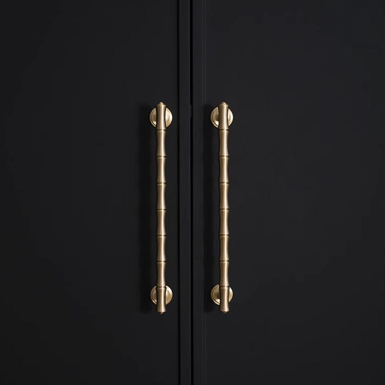 May include: Two gold-coloured cabinet handles with a bamboo design are mounted on a black cabinet door. The handles are vertically oriented, with a cylindrical bamboo-like texture and rounded ends.