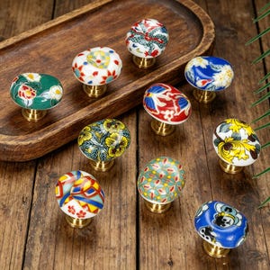Puede incluir: Una colección de tiradores de cerámica decorativos con bases de latón. Los tiradores presentan varios diseños florales y estampados coloridos, incluyendo rojo, azul, amarillo y verde, sobre un fondo blanco. Los tiradores están dispuestos sobre una superficie de madera.